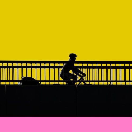 Silhouette of a person riding a bike across a bridge, against a yellow sky and pink foreground.